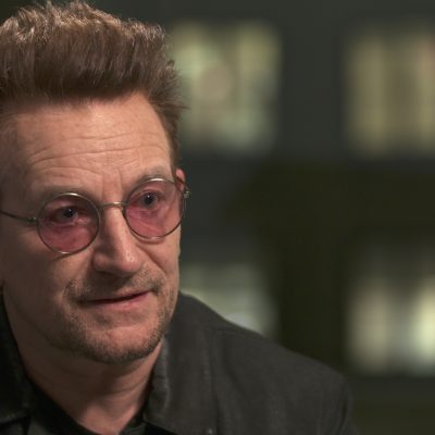 Portrait of singer-songwriter Bono wearing pink-tinted glasses and a suit.