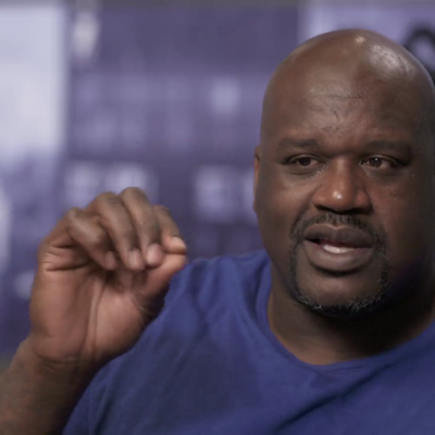 Portrait of Shaquille O'Neal wearing a blue shirt, hands gesturing, and in mid-speech.