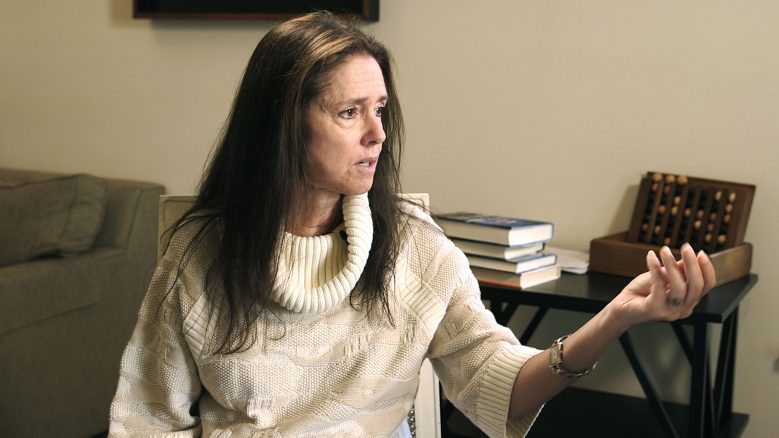 Portrait of Julie Taymor wearing a white top, in mid-speech, and left hand outstretched.