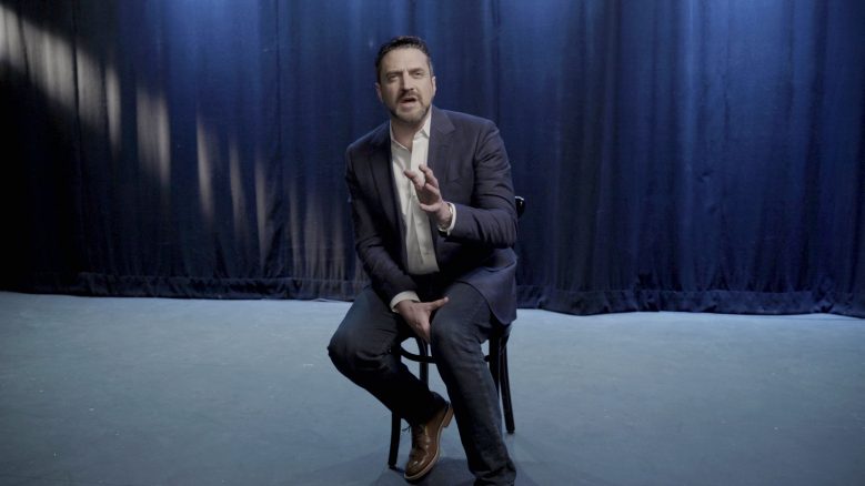 Full body shot of Raúl Esparza seated on a chair, gesturing with his hands and in the middle of speaking.