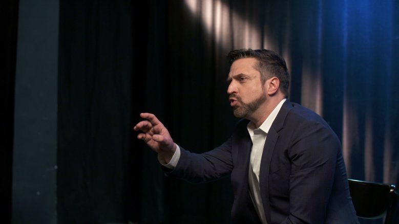 Side view of Raúl Esparza with his eyebrows furrowed and speaking with a hand outstretched.