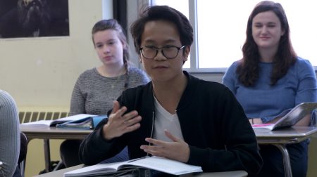 Student seated at a desk, gesturing with hands while speaking.