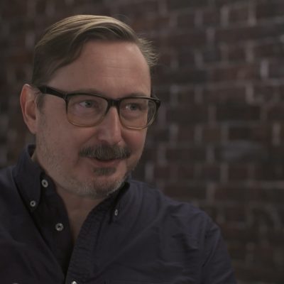 Portrait of John Hodgman wearing a button-up shirt and smiling.