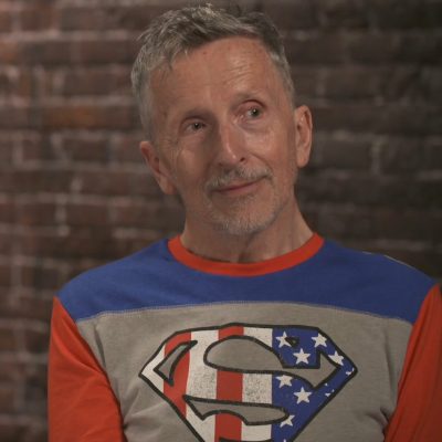 Portrait of Simon Doonan smiling while glancing to the side and wearing a Superman logo shirt.