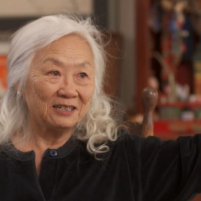 Portrait of Maxine Hong Kingston wearing a black top and in mid-speech with hand gesturing.