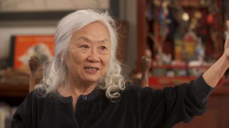Portrait of Maxine Hong Kingston wearing a black top and in mid-speech with hand gesturing.