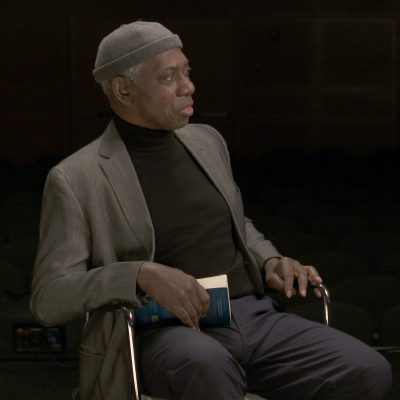 Portrait of Yusef Komunyakaa in a suit, looking to the side, hand holding a book.