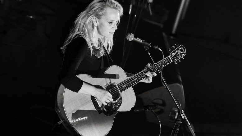 Mary Chapin Carpenter seated with a guitar in her lap, facing a microphone stand.