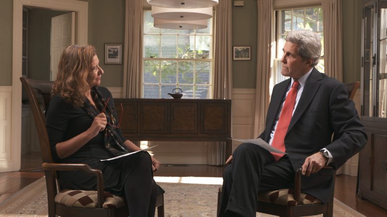 Elisa New and John Kerry seated facing each other, engaged in conversation.