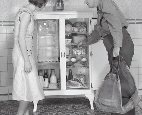 A man and woman standing by a 1920s ice box, man reaching inside to retrieve an item.