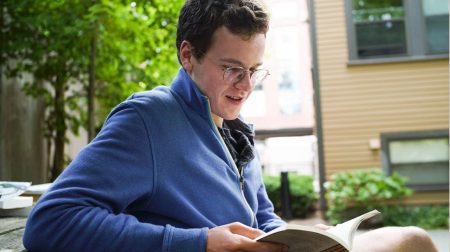 Frank Cahill seated, leaning back on elbows against a table, looking down and reading a book.