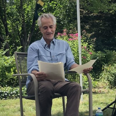 David Strathairn, wearing a blue collared shirt, sits and reads from a pieces of paper.