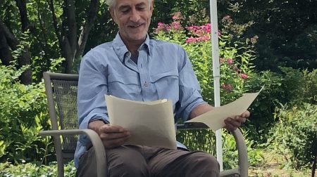 David Strathairn, wearing a blue collared shirt, sits and reads from a pieces of paper.