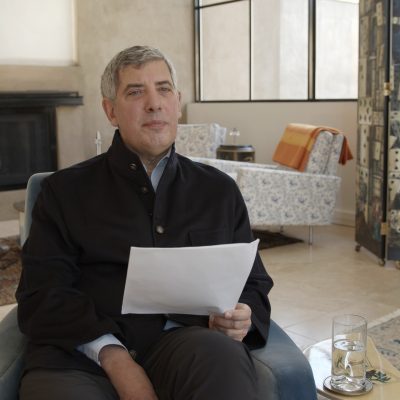 Philip Galanes, sitting in a chair, wears a black jacket and holds a piece of paper in his left hand.