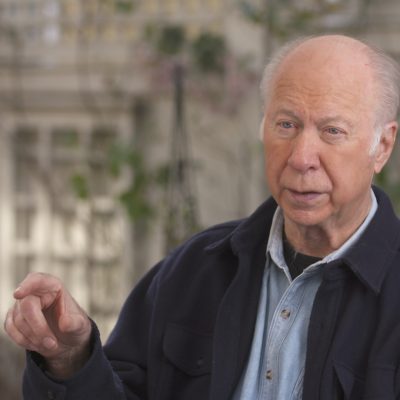 David Gergenw wears a suit while in the middle of talking and gesturing with his hand.