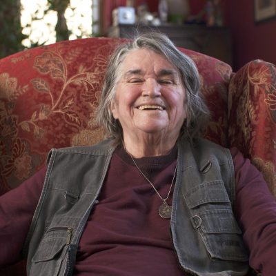 Bernadette Mayer, sitting in a chair, leans back and smiles.