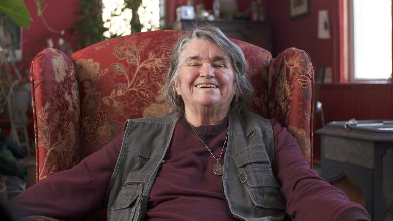 Bernadette Mayer, sitting in a chair, leans back and smiles.