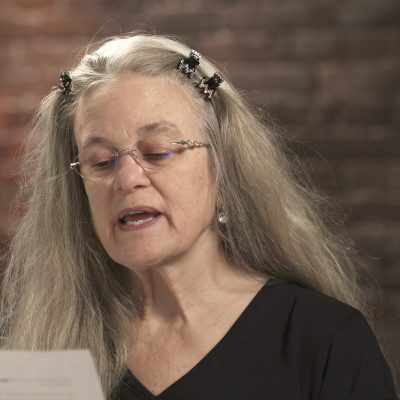 Sharon Olds wears a black top while in the middle of reading from a piece of paper.