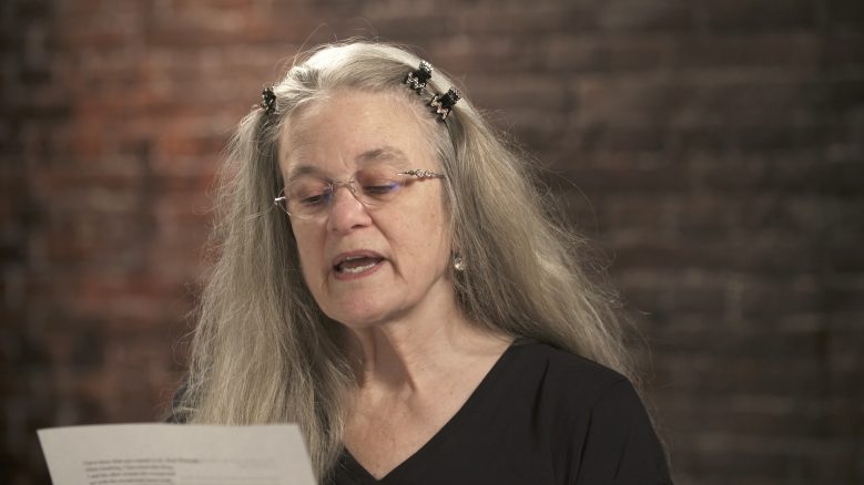 Sharon Olds wears a black top while in the middle of reading from a piece of paper.