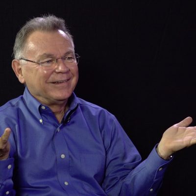 Alberto Rios, wearing a blue collared shirt, smiles while gesturing with his hands.