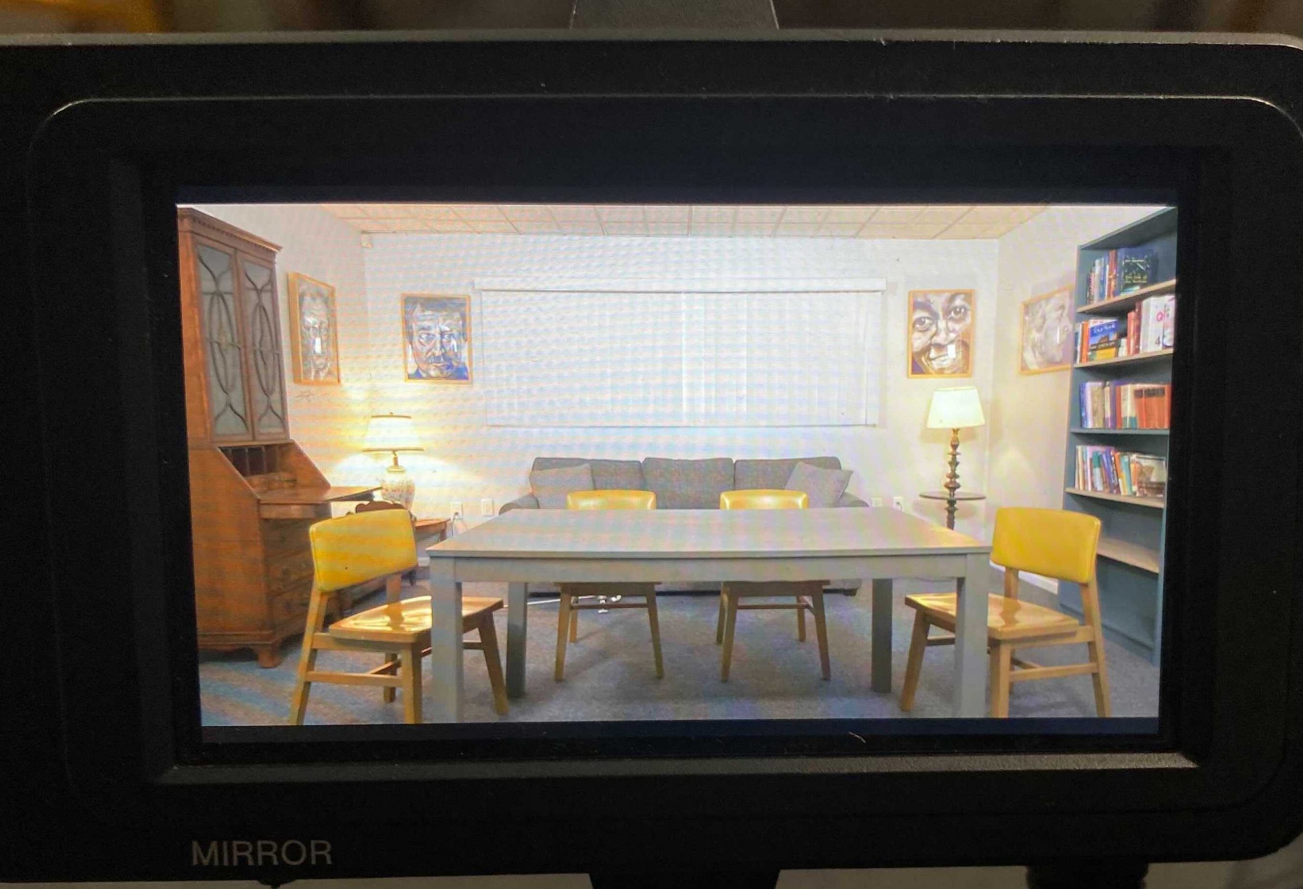 Poetry in America's Brighton, MA office, which has a gray table surrounded by four yellow chairs in the foreground and a sofa with two lamps on either side in the background.