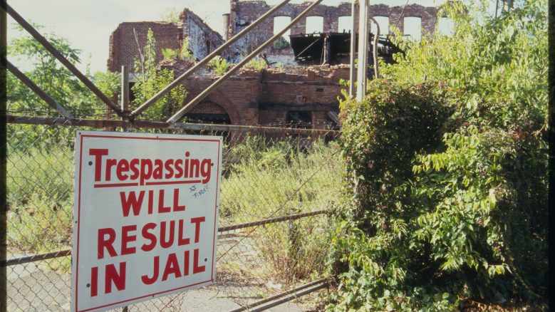 Ruins of Colt factory; sign in foreground reads: "Trespassing will result in jail."