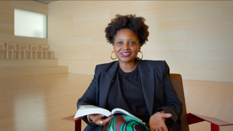 Poet Tracy K. Smith, wearing a black suit, holds a book in her hand while in the middle of speaking.