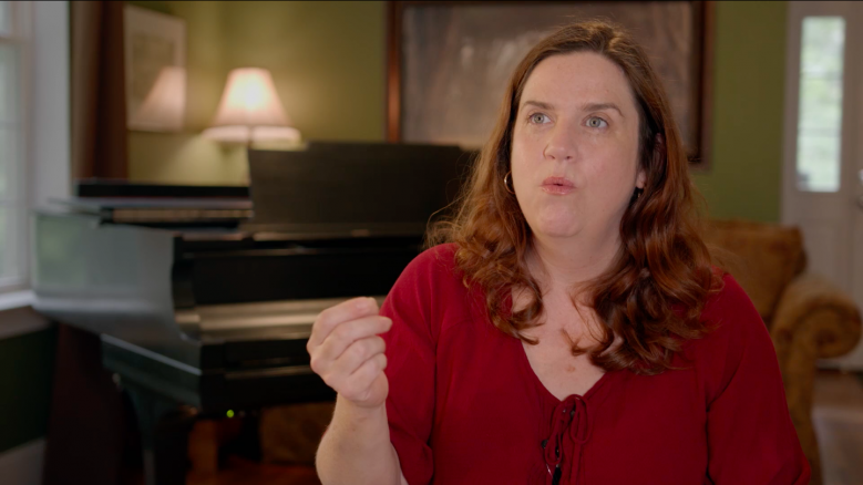 Actor Donna Lynne Champlin, wearing a red blouse, in the middle of speaking while gesturing with her hand.