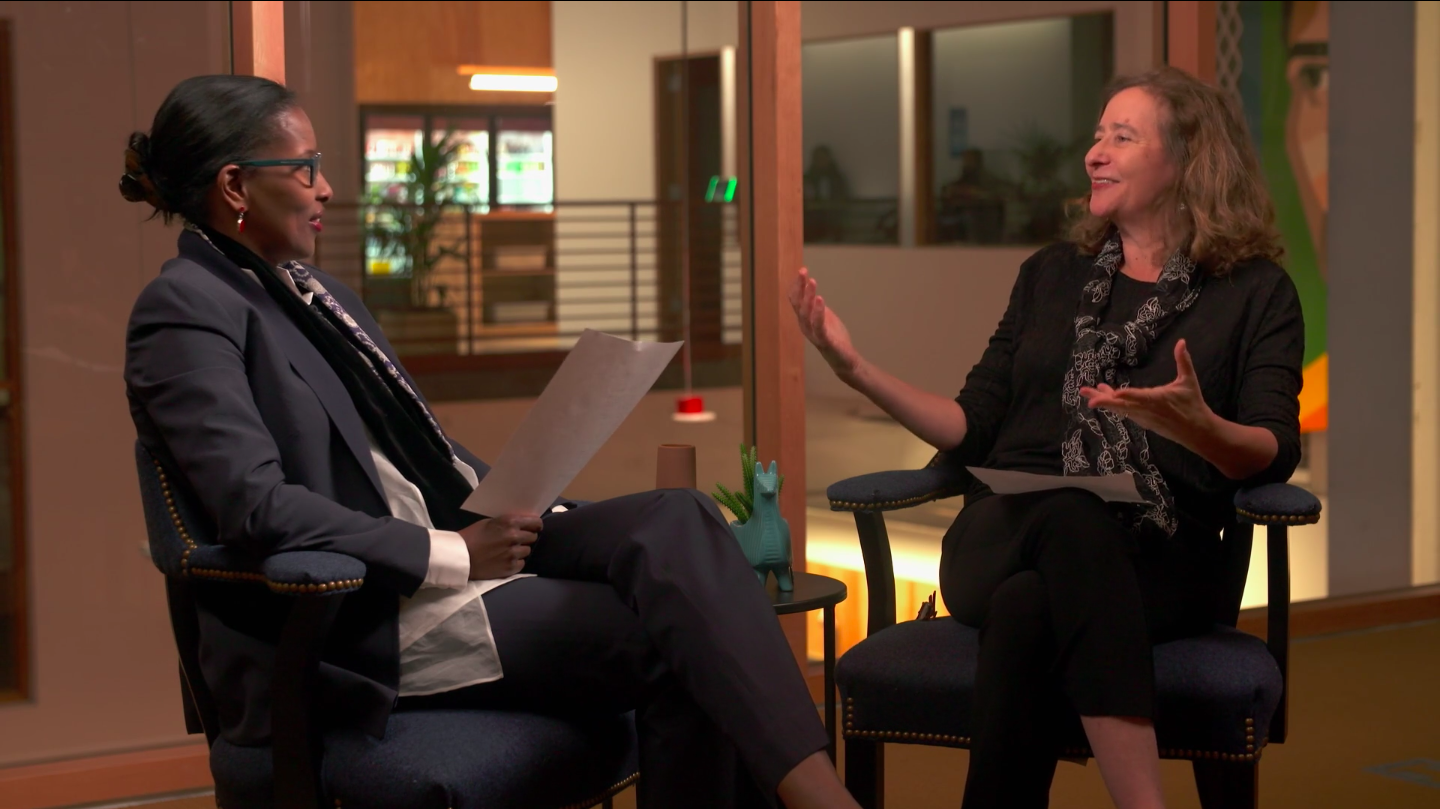 Activist Ayaan Hirsi Ali and host Elisa New sit facing each other. Elisa is gesturing while in the middle of speaking, and Ayaan holds a paper while looking at Elisa.