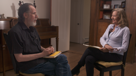 Martín Espada, wearing a black collared shirt, sits facing the host, Dr. Elisa New, who is smiling and wearing a blue long-sleeved collared shirt.