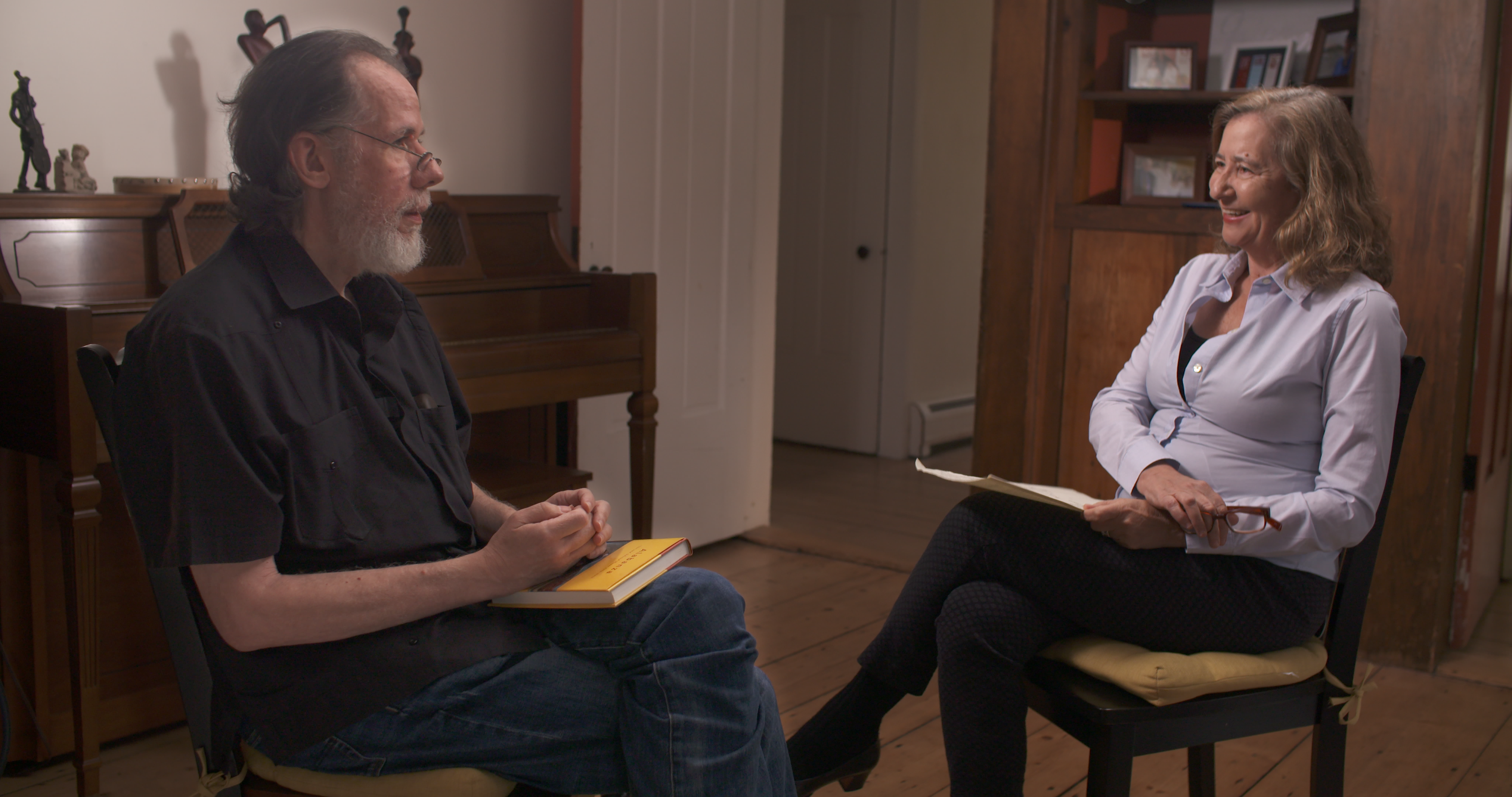 Martín Espada, wearing a black collared shirt, sits facing the host, Dr. Elisa New, who is smiling and wearing a blue long-sleeved collared shirt.