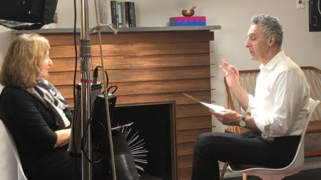 A behind the scenes of host Elisa New and actor John Turturro facing each other while in the middle of speaking, surrounded by cameras and recording equipment