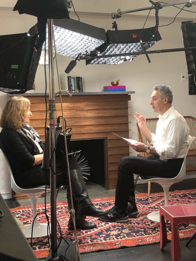 A behind the scenes of host Elisa New and actor John Turturro facing each other while in the middle of speaking, surrounded by cameras and recording equipment