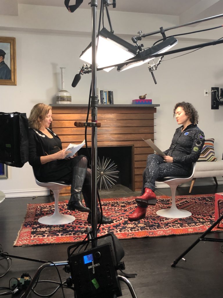 A behind the scenes of host Elisa New and writer Maria Popova facing each other while in the middle of reading papers from their hands, surrounded by cameras and recording equipment.