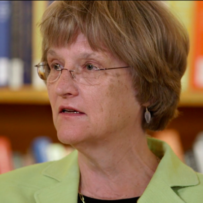 Portrait of Drew Faust wearing a green blazer and looking the left, in the middle of speaking.