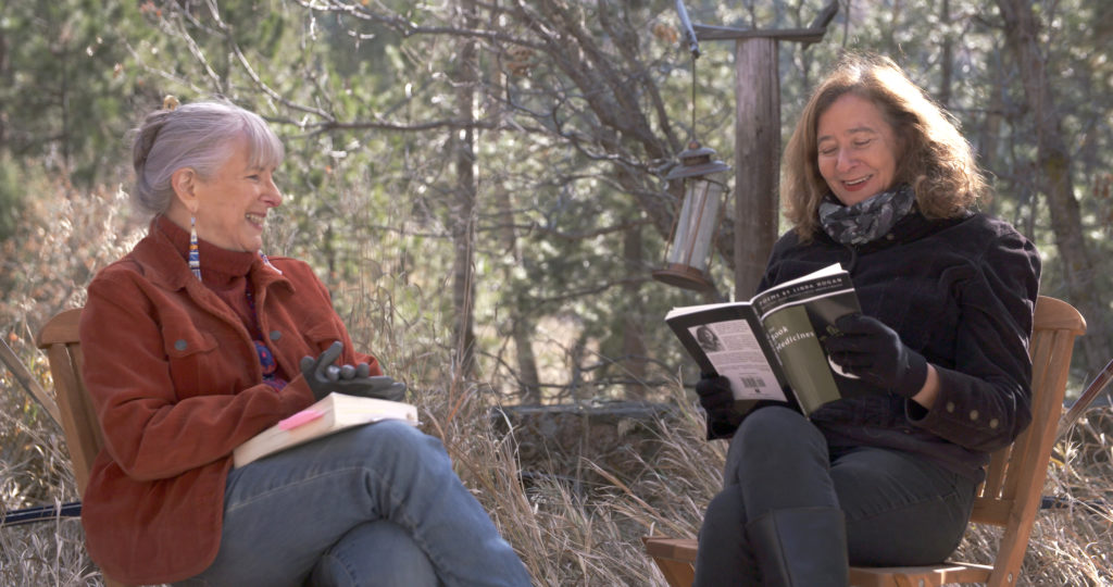 Featured poet Linda Hogan, wearing a brown jacket, smiles while sitting facing Professor Elisa New, who is wearing a black jacket and reading from a book.