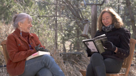 Featured poet Linda Hogan, wearing a brown jacket, smiles while sitting facing Professor Elisa New, who is wearing a black jacket and reading from a book.