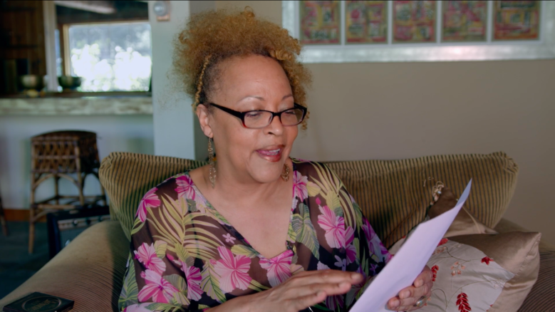 Cassandra Wilson wearing a floral blouse, in the middle of reading from a paper in her hands