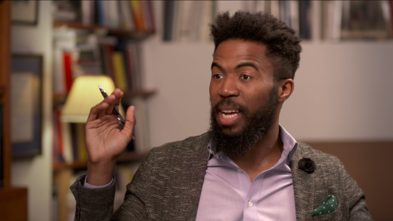 Dr. Joshua Bennett, wearing a suit, in the middle of speaking while gesturing with his right hand.