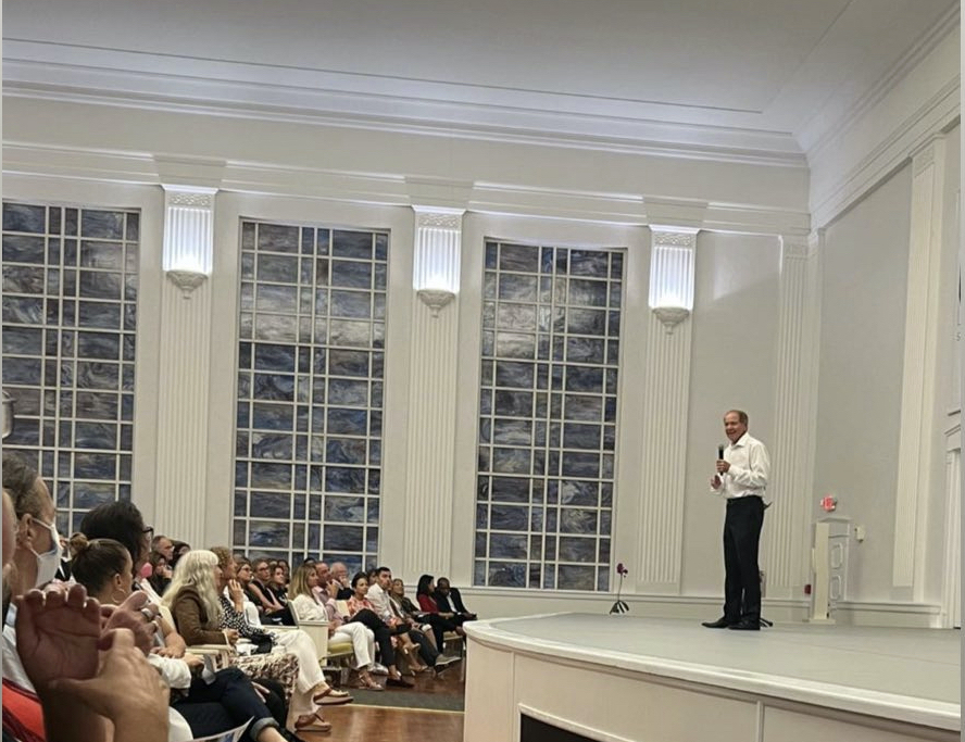 A speaker holding a microphone and wearing a white-collared shirt and black slacks stands on top of a stage overlooking a crowd, introducing Richard Blanco at his event.