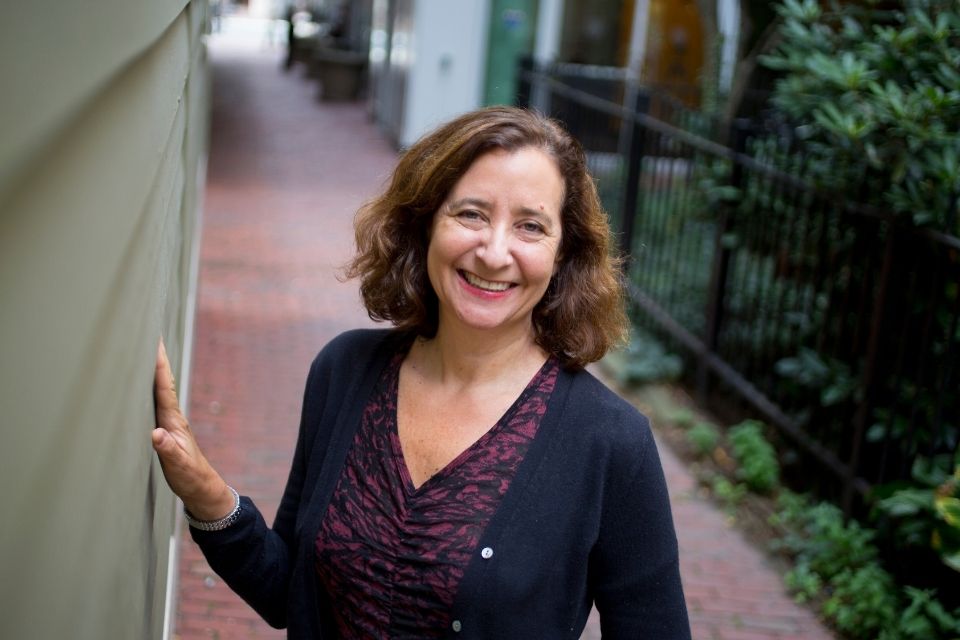 Elisa New wears a purple blouse and navy cardigan and smiles as she is pictured outside.