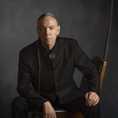 Jerod Tate wears a black suit and shirt, sitting on a wooden chair in front of a grey backdrop.