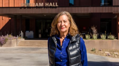 Elisa New stands front of Durham Hall, wearing a blue shirt and a black vest.