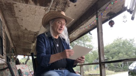 Jimmy Dale Gilmore wears boots, jeans and a wide-brim hat as he sits on a stool on a porch and reads from the paper in his hands.