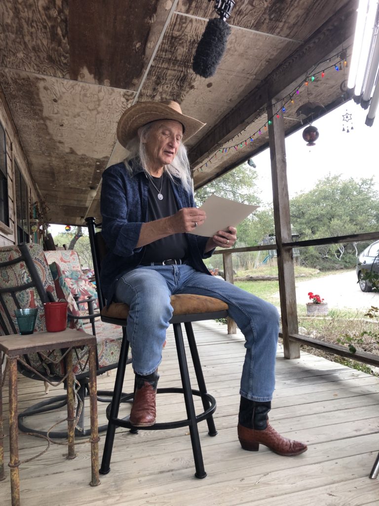 Jimmy Dale Gilmore wears boots, jeans and a wide-brim hat as he sits on a stool on a porch and reads from the paper in his hands.
