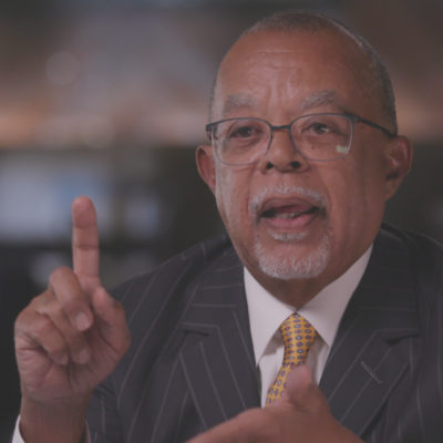 Henry Louis Gates wears glasses and a suit with a yellow tie, and raises a fingers as he speaks.