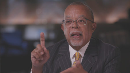 Henry Louis Gates wears glasses and a suit with a yellow tie, and raises a fingers as he speaks.