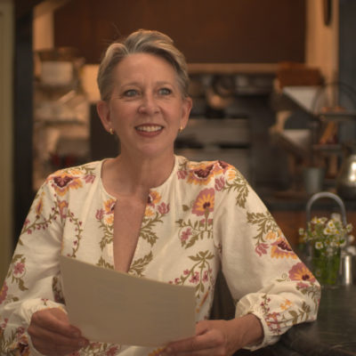 Gabrielle Hamilton wears a flowery top and smiles, reading from a piece of paper in her kitchen.