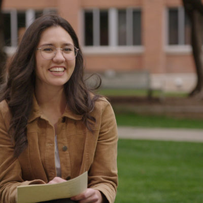 Mikayla Mace Kelley smiles and wears a brown shirt, as she holds some papers whilst sitting in a grassy courtyard.