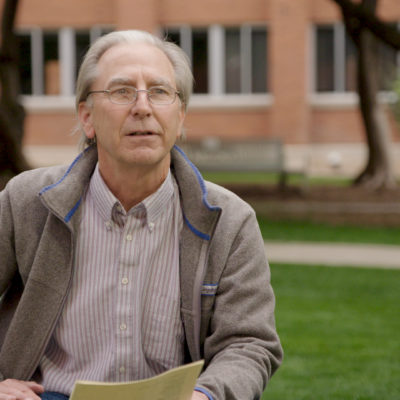 Dr Barry Pryor wears a grey shirt and fleece, as he sits outside in a grassy courtyard.
